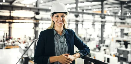 Frau mit Schutzhelm und Blazer steht lächelnd in moderner Produktionshalle.
